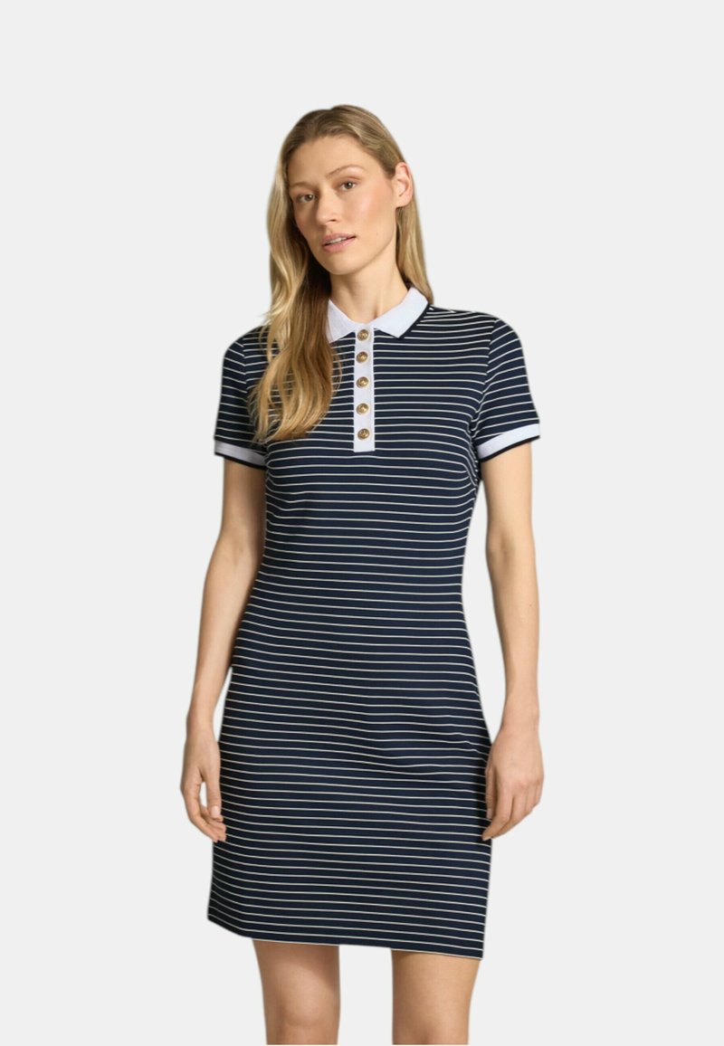 Femme portant une robe polo à manches courtes rayée bleu marine et blanche avec un col blanc et des boutons dorés, debout devant un fond uni.