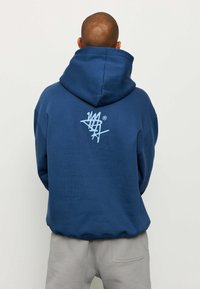 Felpa con cappuccio blu navy realizzata in tessuto morbido. Presenta una grande grafica azzurro chiaro sul retro e una vestibilità ampia e rilassata.