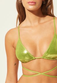 Groene metallic driehoek bikini-top met dunne verstelbare bandjes en een kruisachtige achterkant. Glad oppervlakte met een glanzende afwerking en minimale bedekking.