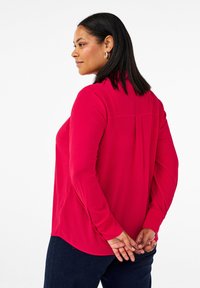 Garā piedurkņu blūze spilgti sarkanā krāsā ar apaļu apakšmalu, aizmugures plisēšanu un mīkstu, gludu auduma tekstūru.