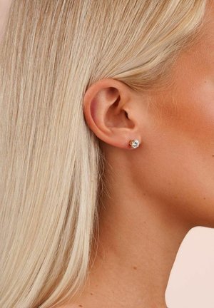 Guldtonad studörhänge med en klar, rund kristall i mitten, mot lätt rufsig blont hår, som visar en elegant glans.