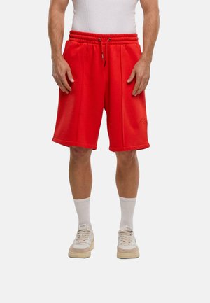 Rote Baumwollshorts mit elastischem Bund und Kordelzug, ausgestattet mit Seitentaschen und einem schlichten Logo. Getragen mit weißen Socken und Turnschuhen.