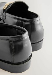 Mocassins en cuir noir avec une finition brillante, présentant un bord cousu et un petit talon. Inclut un accent doré sur le dessus.