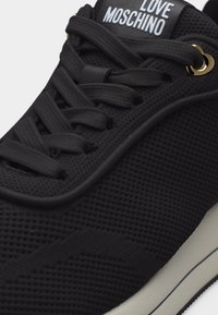 Μαύρο αθλητικό παπούτσι από δίχτυ με μαύρα κορδόνια, λευκό λογότυπο "LOVE MOSCHINO" στη γλώσσα και χρυσές λεπτομέρειες στα κορδόνια κοντά στην κορυφή.