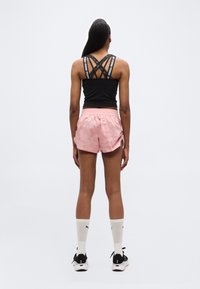 Γυναίκα στέκεται με την πλάτη, φοράει μαύρο αθλητικό τοπ με λουριά, ροζ αθλητικό σορτς, λευκές κάλτσες Puma και μαύρα αθλητικά σε απλό φόντο.