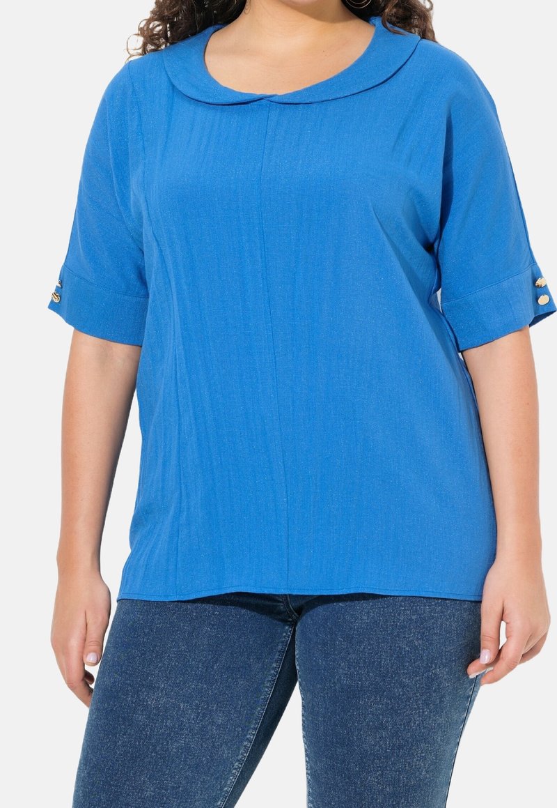 Femme portant un top bleu à manches courtes avec des boutons dorés sur les manches et un jean en denim bleu foncé coupé au niveau de la mi-cuisse.