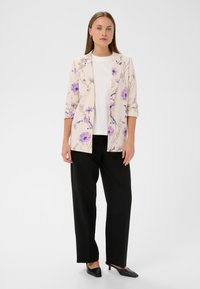 Blazer floral en beige clair avec des accents violets, porté sur une chemise blanche, associé à un pantalon noir et des escarpins noirs pointus.