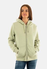 Veste à capuche zippée vert clair en tissu doux, avec une poche frontale, un cordon de serrage à la capuche et des poignets côtelés. Le modèle se tient avec les mains dans les poches.