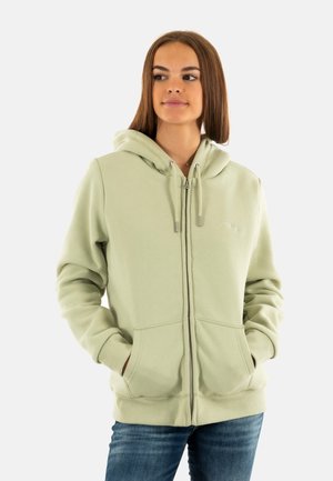 Veste à capuche zippée vert clair en tissu doux, avec une poche frontale, un cordon de serrage à la capuche et des poignets côtelés. Le modèle se tient avec les mains dans les poches.