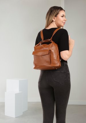 Vrouw met een zwart shirt en donkere jeans, draagt een bruine leren rugzak met een voorvak, staand in een minimalistische kamer met witte blokken.