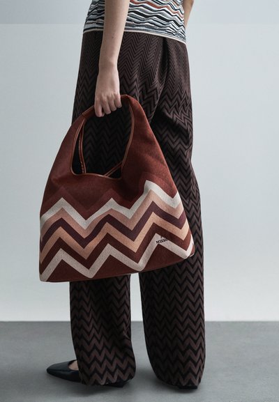 Άτομο που κρατά μεγάλη καφέ τσάντα με ζιγκ-ζαγκ σχέδιο, φορώντας ταυτόχρονα παντελόνι σε ταιριαστό καφέ ζιγκ-ζαγκ σχέδιο και μαύρες μυτερές γόβες.