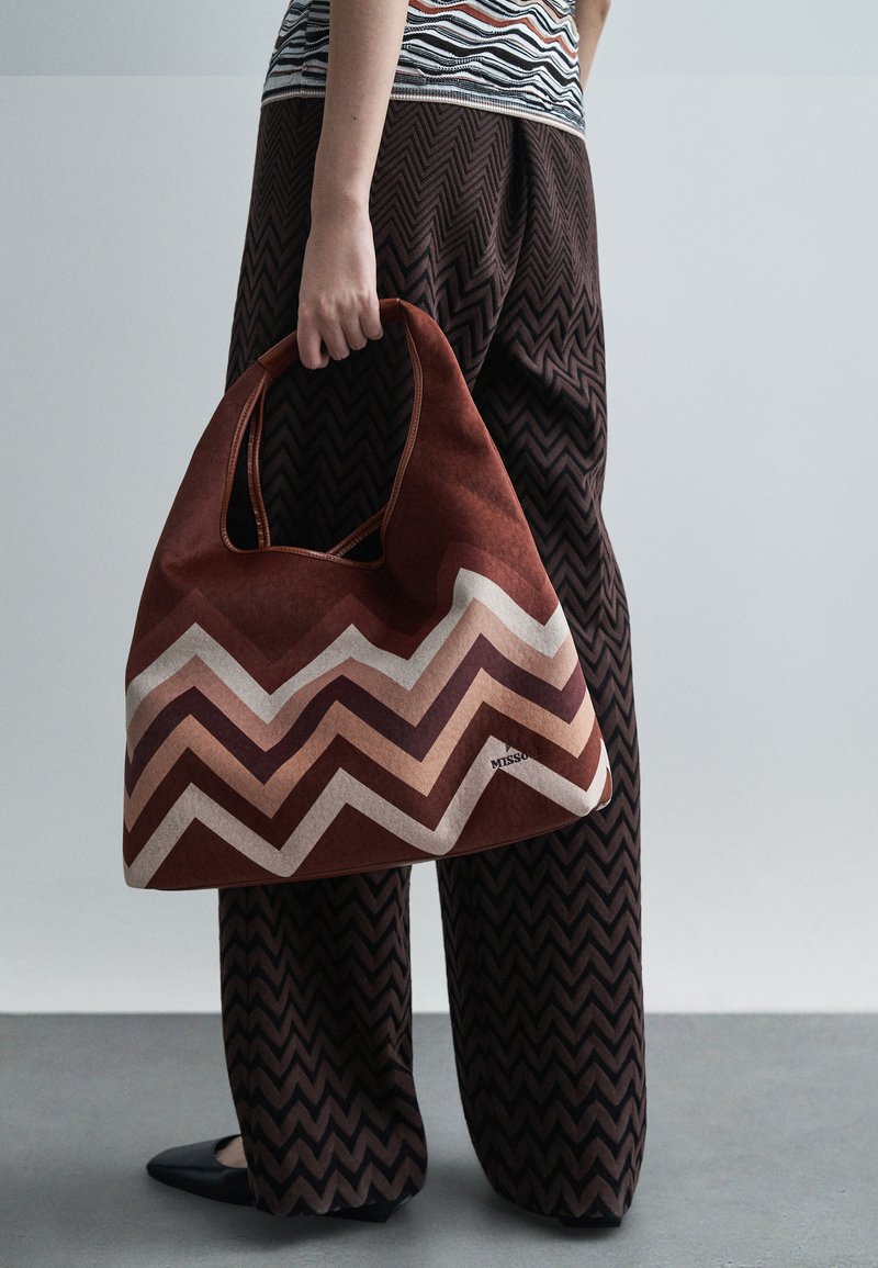 Personne tenant un grand sac à main marron avec un motif en zigzag, portant un pantalon marron à motif en zigzag assorti et des chaussures noires à talons pointus.