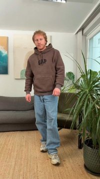 Brauner Fleece-Hoodie mit vorderer Tasche und schwarzem "GAP"-Logo, blaue Jeans und beige Sneaker, stehend neben einer Topfpflanze.
