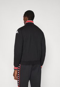 Svart jacka med hög krage, med röda och vita randiga detaljer. Slät textur, åtsittande design, med logotypdetalj på axeln.