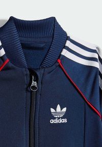 Mornarsko modra adidas jakna na zadrgo z rebrastim ovratnikom, belimi trakovnimi rameni, rdečimi akcenti in belim logotipom na prsih.
