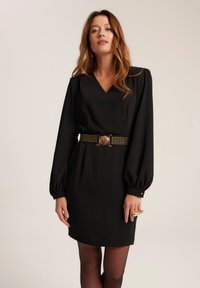 Czarna sukienka z dekoltem w serek, długimi bufkami i dopasowanym krojem. Podkreślona czarnym paskiem z goldowymi ćwiekami i okrągłą klamrą.