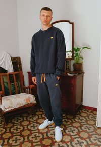Navy sweatshirt and sweatpants with orange drawstring, featuring a small elephant logo. Model wears white sneakers, standing on patterned tile floor.