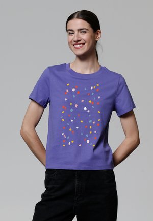 T-shirt violet à manches courtes en coton, présentant un motif éclaboussé coloré et abstrait dans des teintes de rouge, jaune, bleu et blanc.