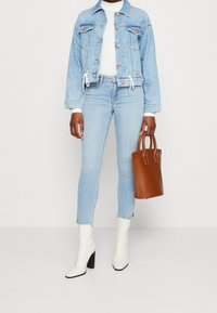 Chaqueta de mezclilla azul claro con cordón, camisa blanca de manga larga, jeans claros, botas de tacón blancas y un bolso de mano de cuero marrón.