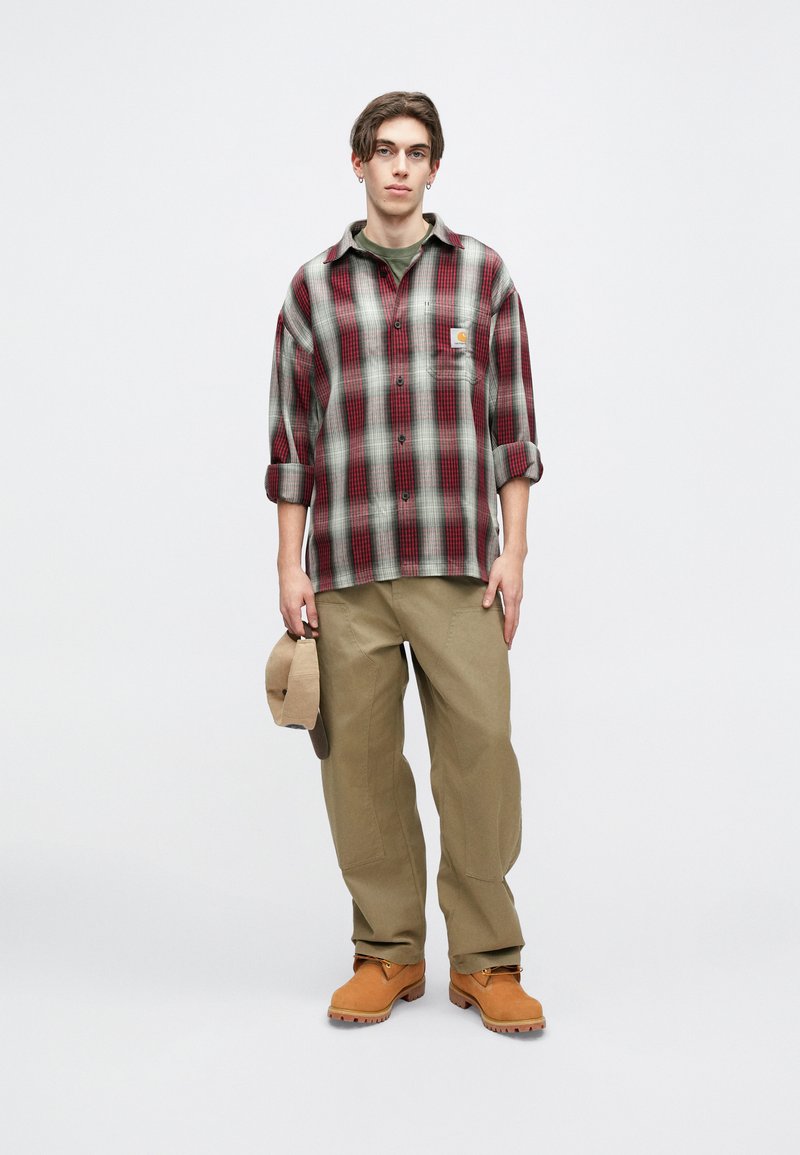Plaid button-up shirt in red and gray, khaki pants, and brown boots. Model holds a beige hat. Textured fabric and relaxed fit.
