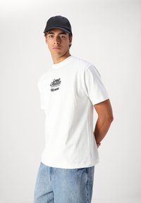 T-shirt en coton blanc avec un logo noir imprimé sur la poitrine. Assorti à un jean en denim bleu clair et une casquette noire.