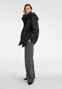 Polstret sort jakke med blomstermønster, med hætte, lynlåslukning og sidelommer. Sammen med grå brede bukser og brune sko.