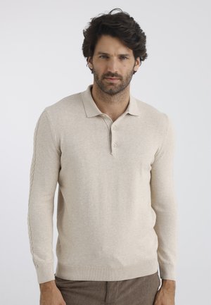 Homme aux cheveux foncés et à la barbe, portant un polo beige à manches longues et un pantalon marron sur un fond gris clair uni.