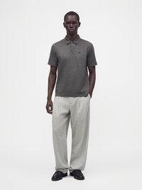 Homme portant un polo à manches courtes gris foncé, un pantalon large gris clair et des chaussures noires, debout sur fond blanc.