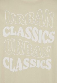 Beige t-krekls ar izteiksmīgu baltu stilizētu tekstu, kas lasāms "URBAN CLASSICS" dažādos izmēros un formās, uz gludas auduma tekstūras.