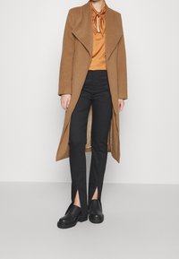 Langer, brauner Mantel mit offenem Vorderteil, kombiniert mit einer orangefarbigen Satinbluse und schwarzen Hosen mit geschlitzten Säumen, ergänzt durch schwarze Schuhe.