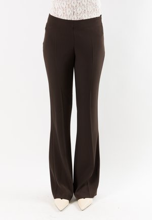 Donna che indossa pantaloni marrone scuro a gamba larga con dettagli a piega, abbinati a scarpe bianche con punta appuntita e top in pizzo.