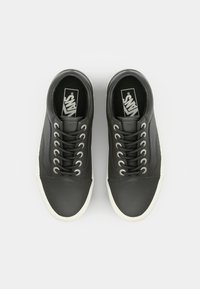 Zapatillas de cuero negras con superficie texturizada, suela de goma blanca y seis ojales metálicos. Presentan un logotipo en la lengüeta y detalles de costura.