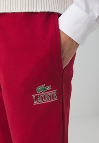 Röda bomullsbyxor med grön krokodil-logotyp och texten "LACOSTE", med ribbad midja och detaljerade sidfickor.