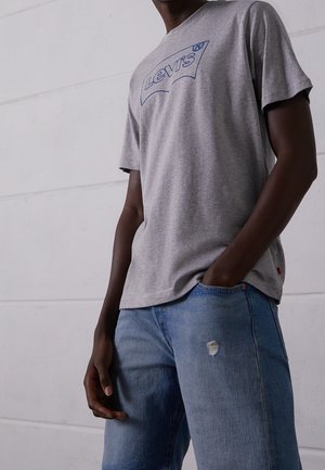 Mężczyzna w szarej koszulce Levi's i jasnoniebieskich dżinsach z małą dziurą na lewym udzie, z ręką w kieszeni, na tle gładkiej białej ściany.