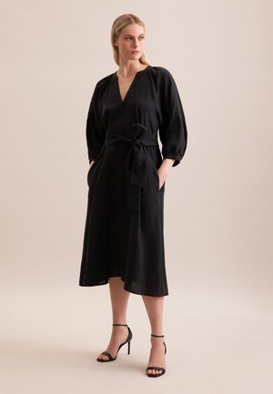 Abito midi nero con lunghe maniche a palloncino, scollo a V profondo e cintura in vita, realizzato in tessuto morbido con tasche laterali, abbinato a tacchi neri con lacci.