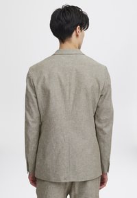 Ljusbrun linneblazer med en texturerad yta, med en enda slits och detaljer med treknappsmuddar, sedd bakifrån.