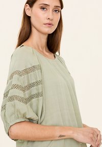 Hellgrüne Bluse mit lockerer Passform, aus strukturiertem Stoff, mit weiten Ärmeln und Spitzenakzenten an den Schultern. Glatte Oberfläche und lässiges Design.