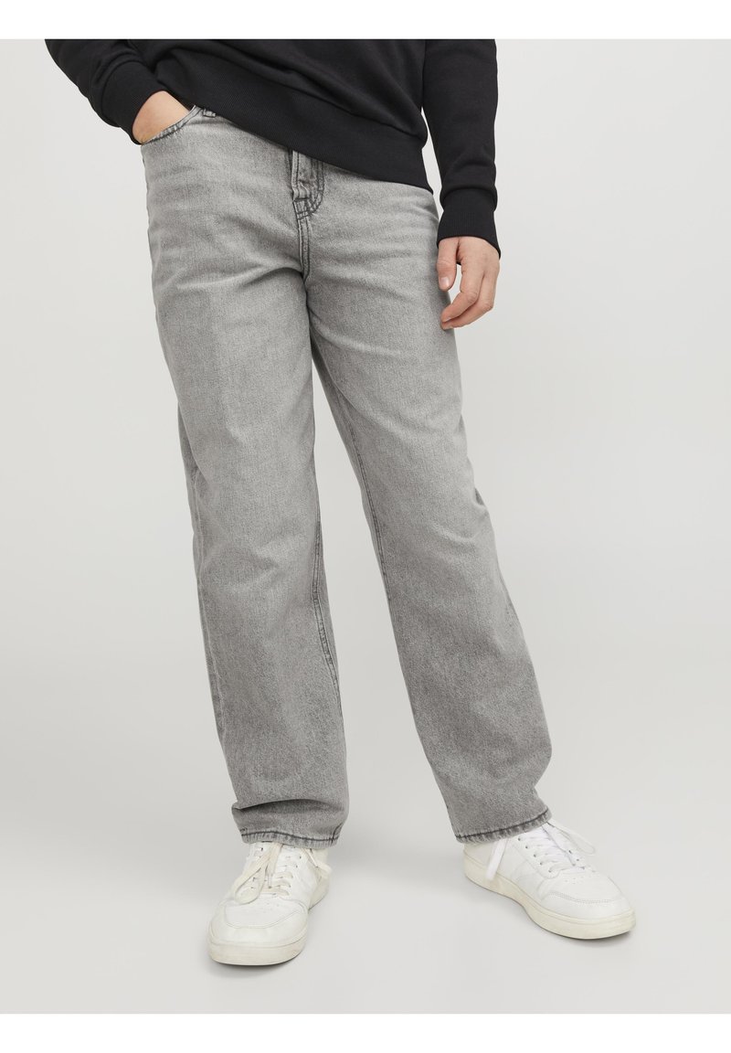 Person som bär ljusgrå rakbenade jeans, vita sneakers och en svart långärmad tröja med en hand i fickan.