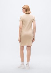 Μπεζ φόρεμα με κοντά μανίκια, εφαρμοστό σχέδιο, μήκος μέχρι το γόνατο και λεία υφή. Φορεμένο με λευκά αθλητικά παπούτσια και κάλτσες αστραγάλου.