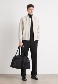 Beež bombersärk lukuga, must kõrge kaelusega särk, mustad püksid ja must logoga spordikott, hele taust.