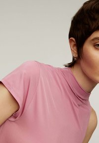 Top ajustado de color rosa claro con cuello alto y mangas cortas, textura suave, suave brillo, y un primer plano de pendientes de cadena plateada y estrellas.