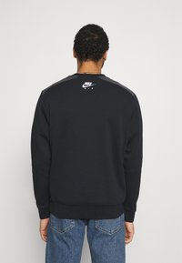 Man i svart Nike Air-tröja och blå jeans, vänd bort mot en enkel ljus bakgrund.