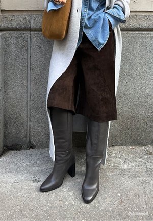 Persoon draagt donkere bruine suéde korte broek, grijze kniehoge leren laarzen, lichtblauw denim overhemd, lange grijze jas en draagt een bruine suéden tas.