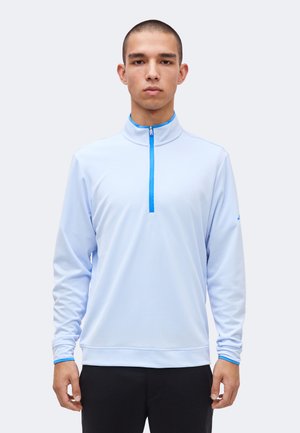 Mand iført en langærmet lyseblå pullover med kvart lynlås og blå kant, stående mod en ensfarvet lys baggrund.