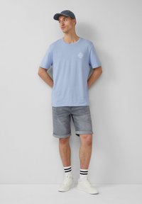 Lichtblauw t-shirt met korte mouwen, grijze denim shorts, witte sneakers en een bijpassende blauwe pet. Verticale strepen op de sokken.