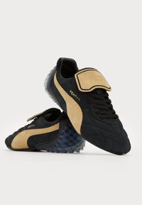 Black and tan sneakers with a textured suede upper, lace closure, padded tongue, and clear bubble sole featuring round spikes. FENTY branding visible.