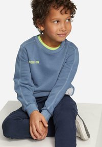 Sweatshirt bleu avec le texte "MOOD ON", poignets côtelés et un col superposé vert néon, associé à un pantalon noir marine et des baskets blanches.