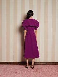 Mulher com cabelo longo e escuro vestindo um vestido midi roxo com mangas bufantes e colarinho em babados, em pé contra uma parede bege listada e um tapete rosa.