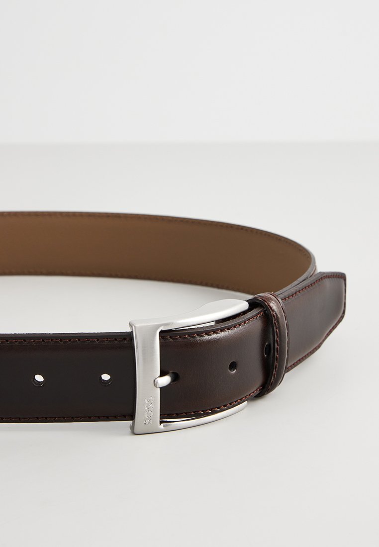 Bruine leren riem met een gladde textuur, voorzien van een zilveren metalen gesp en meerdere gaten voor aanpassing. Eenvoudig ontwerp met contrasterende binnenvoering.