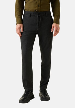 Man draagt donkergrijze broek met trekkoord, zwarte sneakers en een olijfgroen shirt met lange mouwen, staand tegen een witte achtergrond.